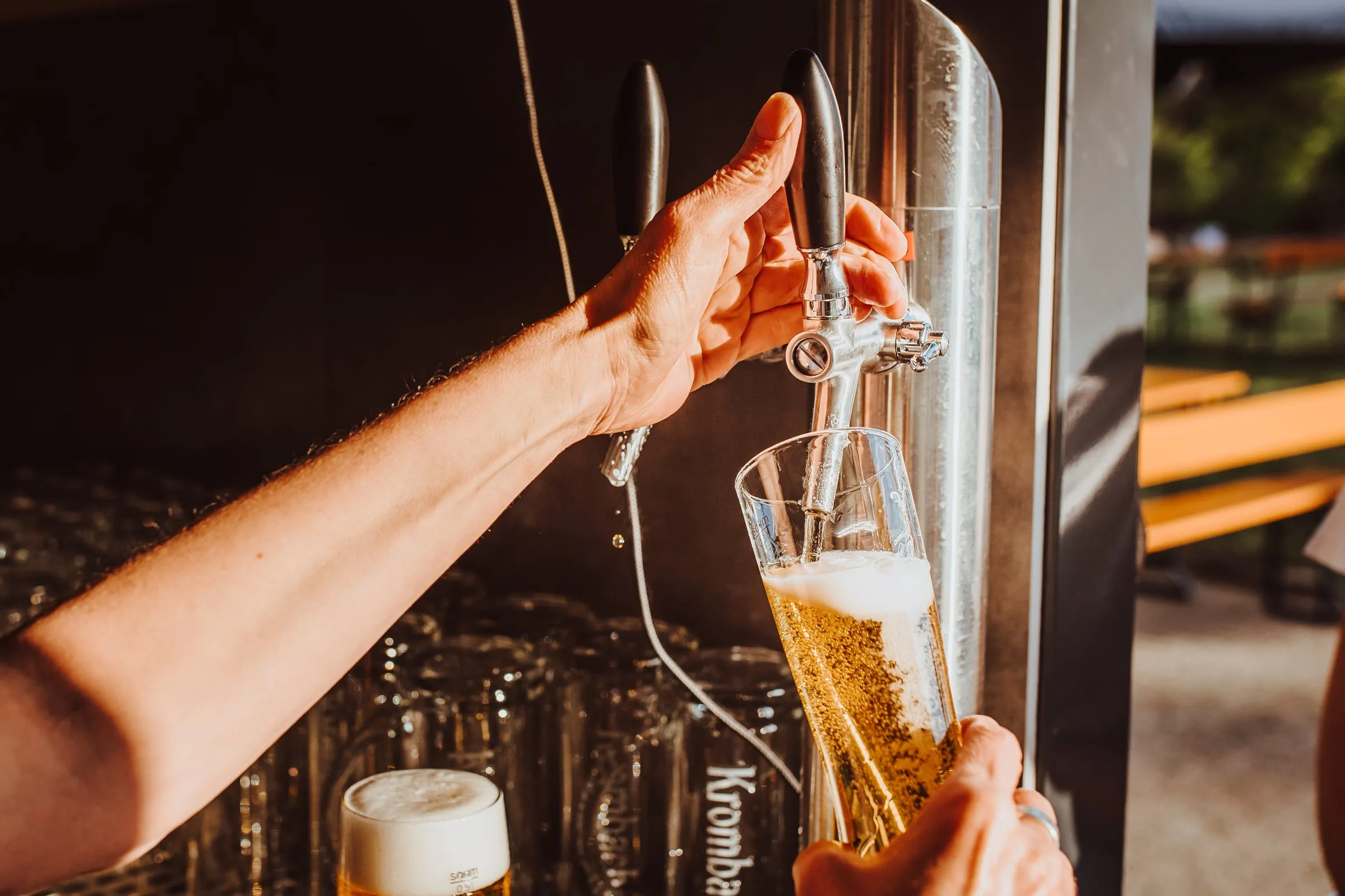 Eine Hand zapft ein frisches Bier am Zapfhahn, im Hintergrund ein Regal mit Gläsern